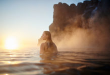 A woman in a hot spring looks at a pink sky.