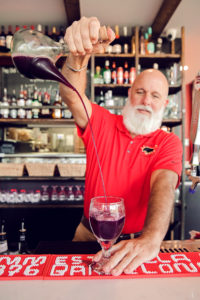 A man with a white beard, in a red polo shirt pours wine in a beautiful restaurant. 