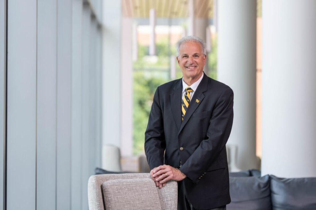 Photo of a man with gray hair in a suit and tie smiling at the camera. He is standing inside resting his hands on the back of a sofa.