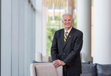 Photo of a man with gray hair in a suit and tie smiling at the camera. He is standing inside resting his hands on the back of a sofa.